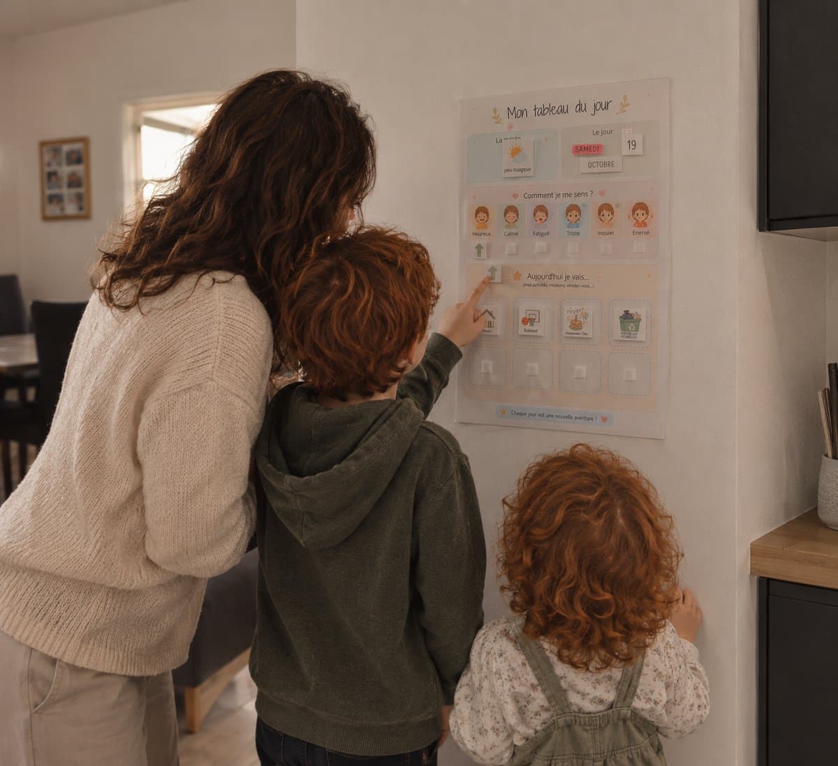 Une maman et ses deux enfants devant le tableau du jour affiché au mur de la cuisine